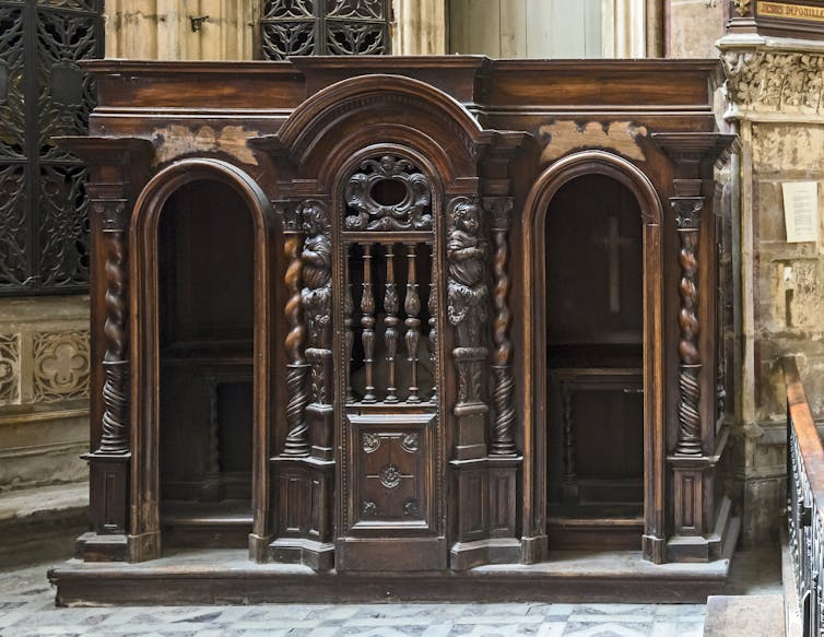 An antique wooden confessional booth with two entrances, and a crucifix inside one of the chambers.
