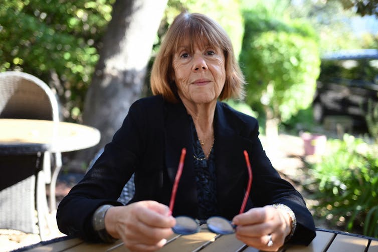 An older woman with short orange hair and bangs looks directly at the camera and holds glasses.