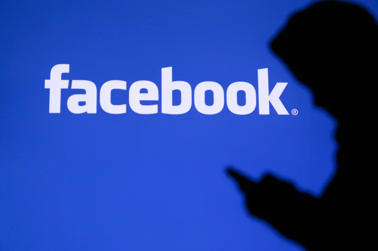 Silhouette of a person wearing a hoodie and looking at a phone against a blue Facebook background.