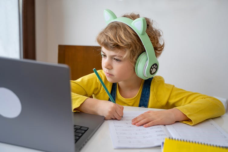 A young child with cat headphones, works on a sheet of paper while looking at a laptop screen.