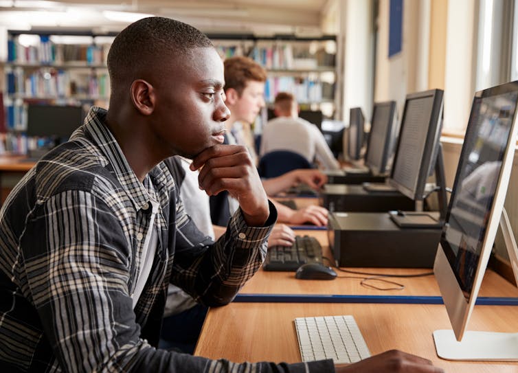 A young man works on a computer in a library. There is a row of desktop computers beside him.