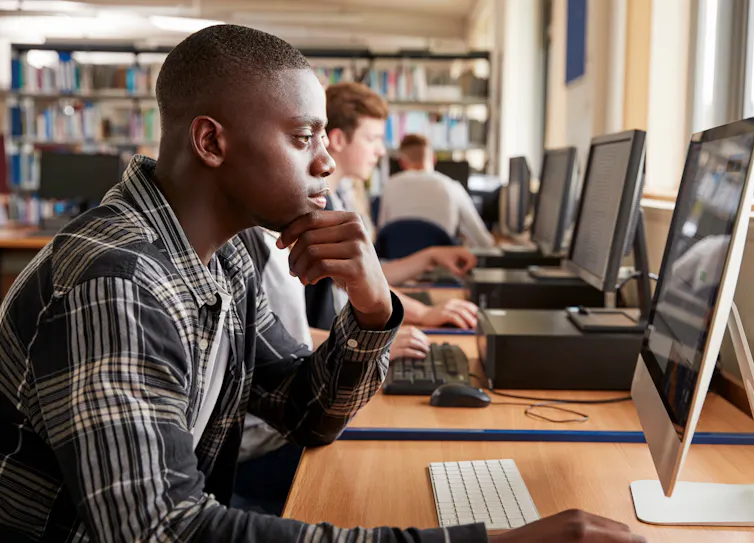 A young man works on a computer in a library. There is a row of desktop computers beside him.