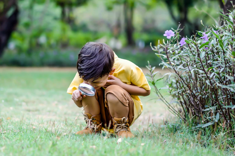 Young boy in yellow shirt and boots looking at grass through a magnifying glass.