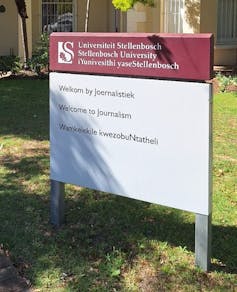 A maroon and white signboard with information in three languages stands before a university building