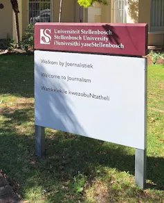 A maroon and white signboard with information in three languages stands before a university building