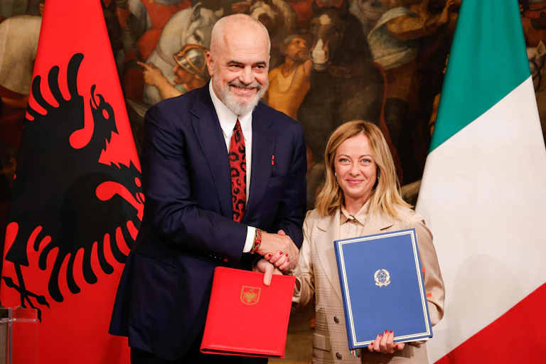 Edi Rama and Giorgia Meloni shake hands while holding diplomatic folders, standing in front of Albanian and Italian flags