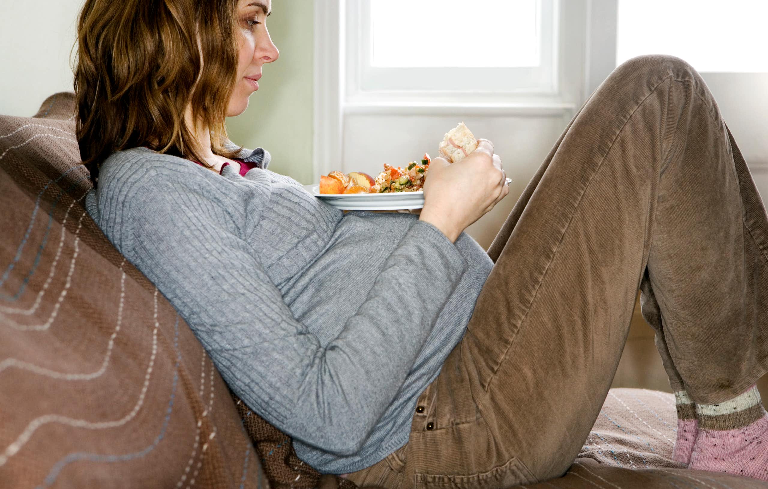 Pregnant woman sitting on couch with a plate of healthy food on her pregnant belly.