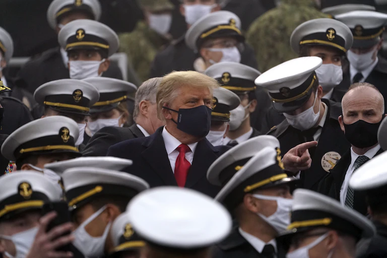 A man with greyish-blond fluffy hair wearing a black face masks stands among uniformed personnel, all wearing white face masks and white rimmed hats.