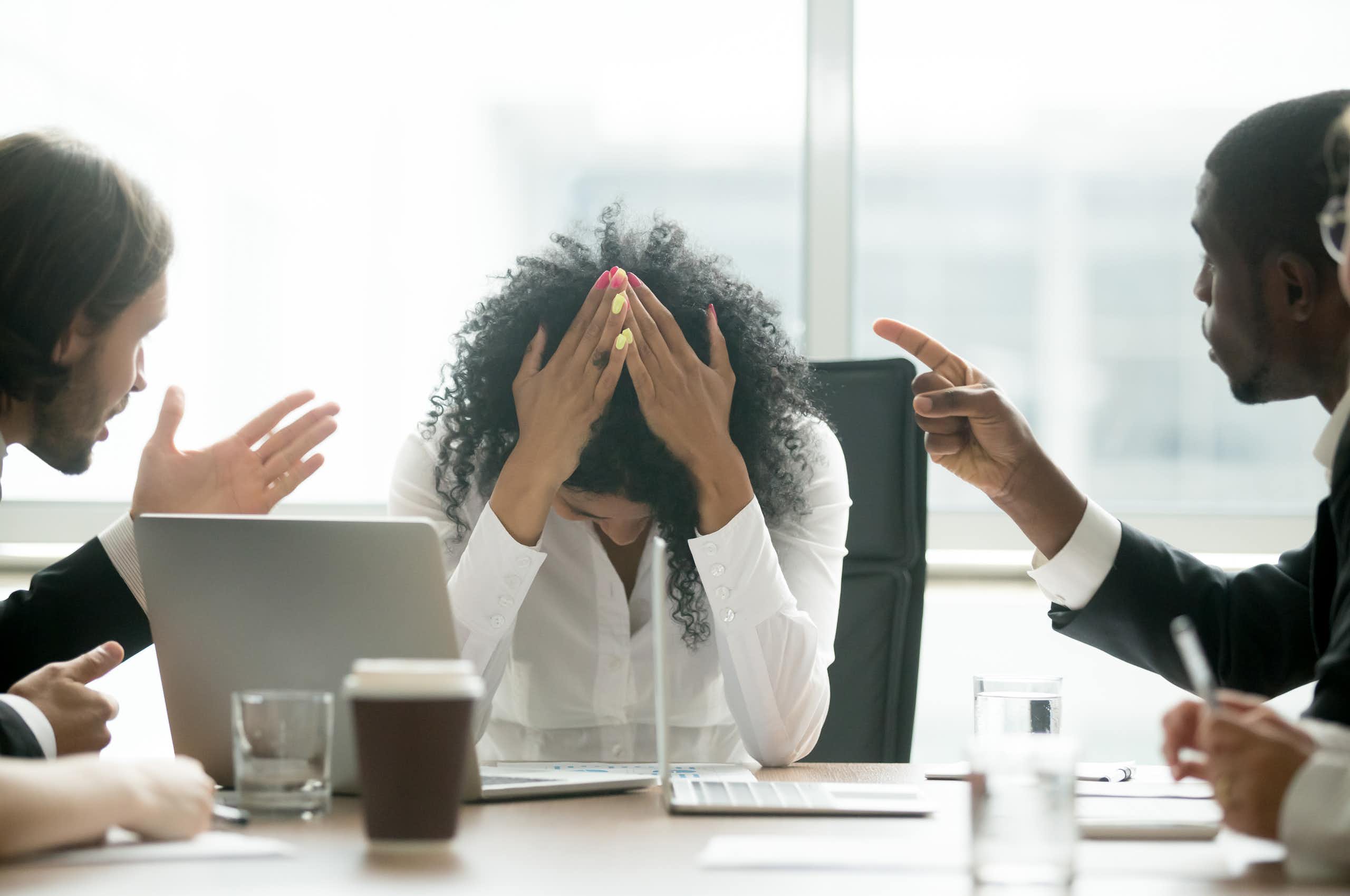 A stressed female professional puts her head in her hands as two leaders berate her.