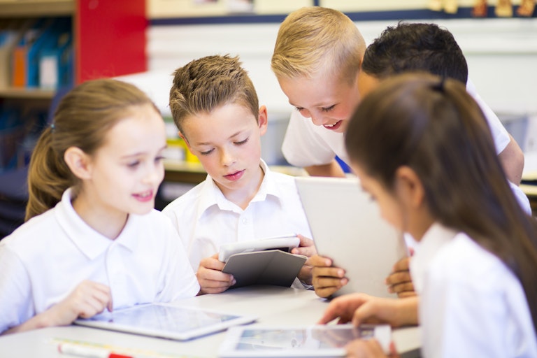 A group of primary students in white uniform shirts look at tablets at a desk.