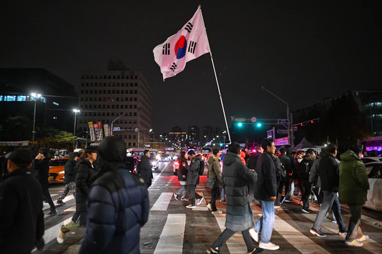 A large crowd of people at cross a street at night. One carries a South Korean flag.