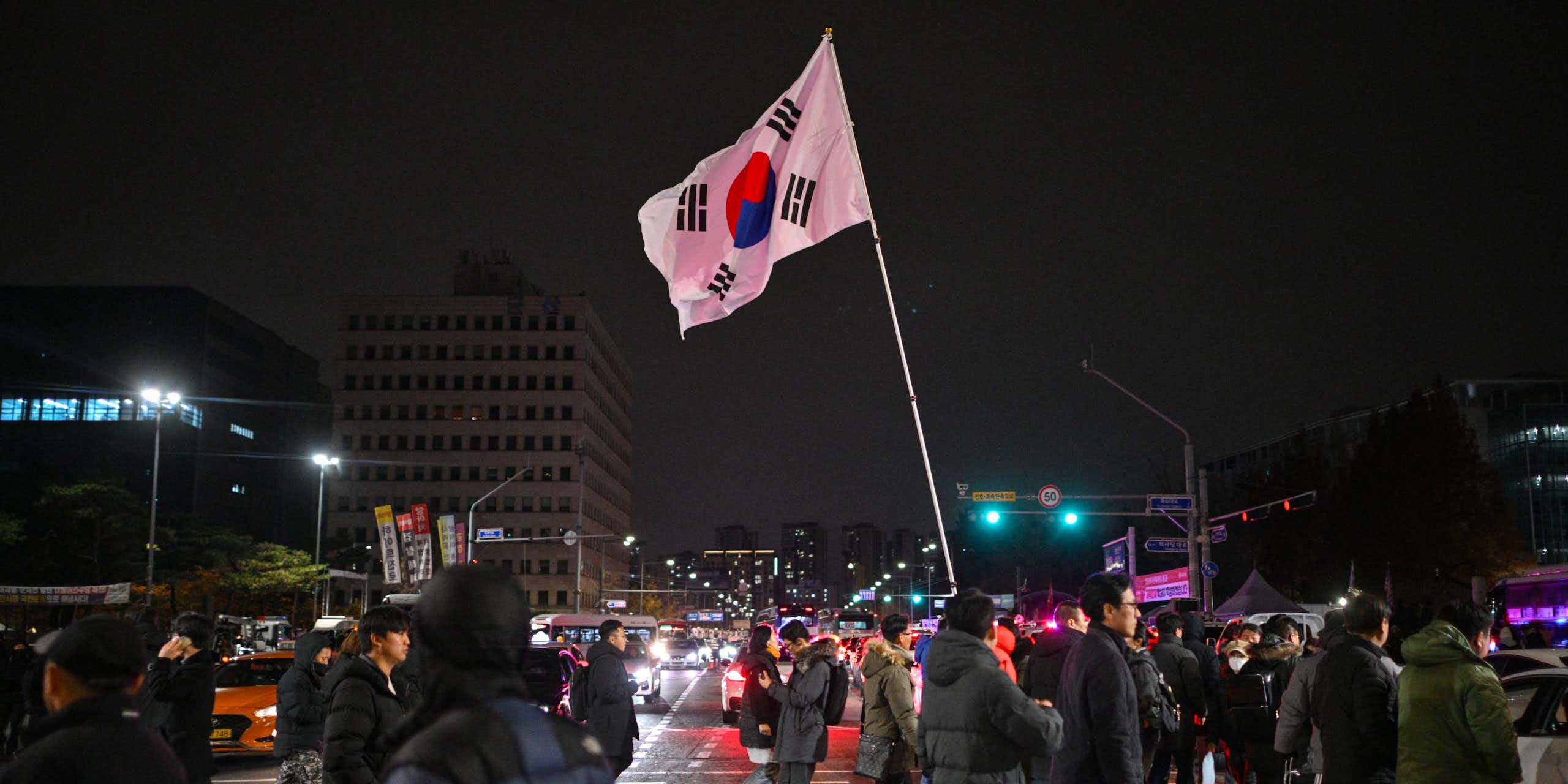 A large crowd of people at cross a street at night. One carries a South Korean flag.