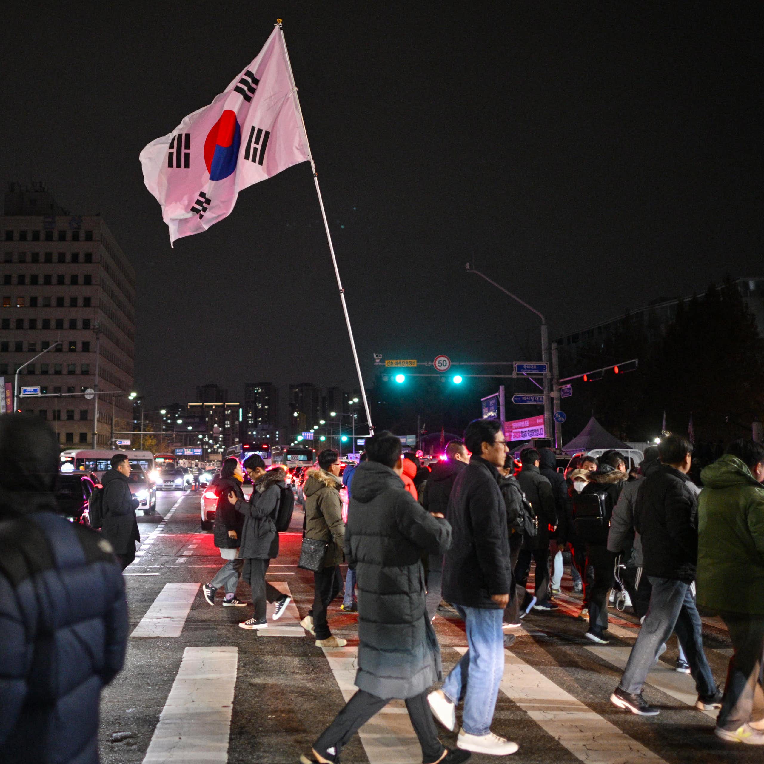 A large crowd of people at cross a street at night. One carries a South Korean flag.