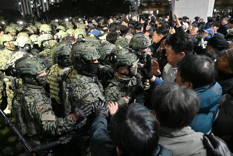 A large crowd of soldiers wearing camouflage and helmets push up against a crowd of civilians.