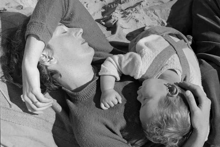 A woman lies on the beach with a young child.