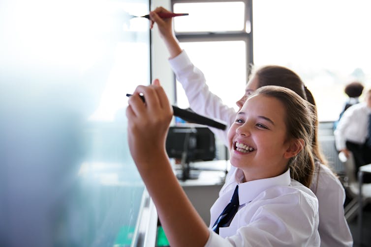Two students write on a white board. One is smiling.