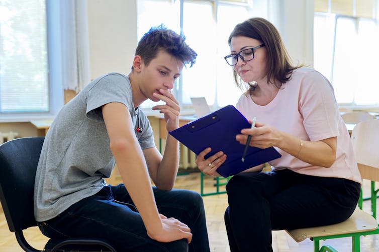 A teenage boy talks to an adult. She is holding a clipboard.
