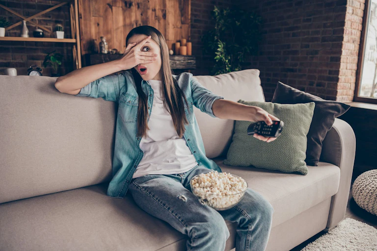 A young woman watches TV with a bowl of popcorn while holding a remote control.