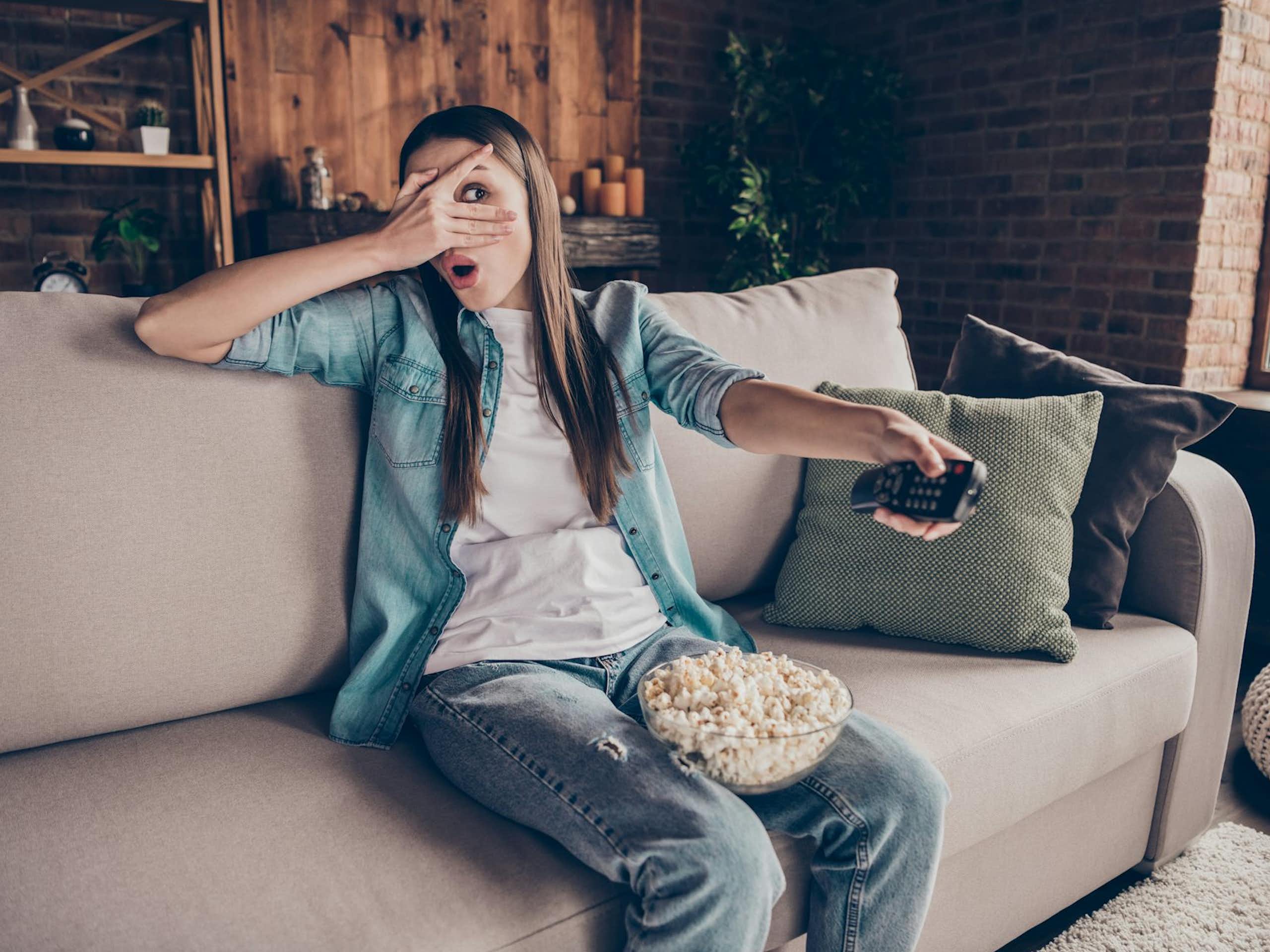 A young woman watches TV with a bowl of popcorn while holding a remote control.