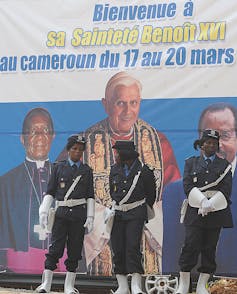 Female soldiers stand in front of a billboard featuring three men, two of them in religious garb.