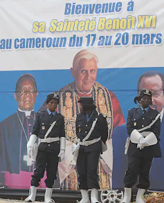 Female soldiers stand in front of a billboard featuring three men, two of them in religious garb.