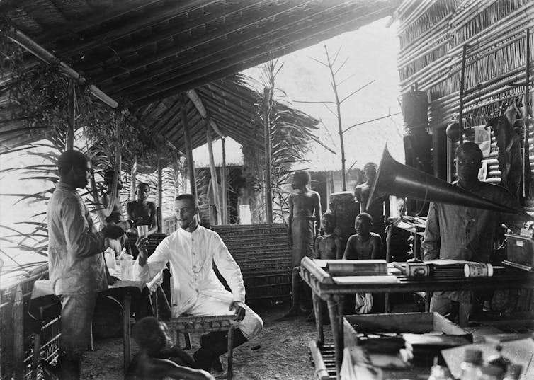 A man in white sits beneath a wooden roof being served by Africans.