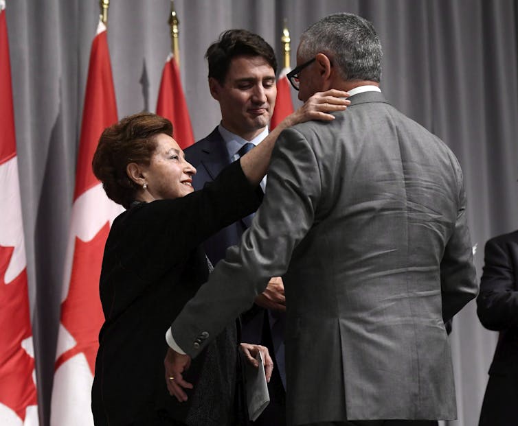 A dark-haired man looks sombre standing next to an older woman who embraces another man.