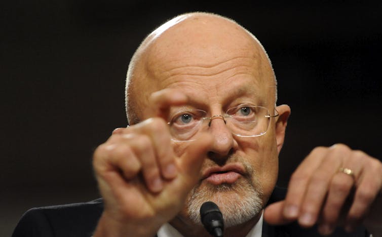 A bald man with a goatee talks into a microphone and gestures with his hands.