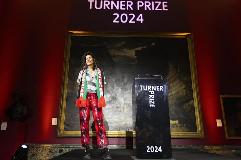 A young Scottish-Asian woman with the Palestinian flag round her neck standing on stage as the Turner Prize winner 2024.