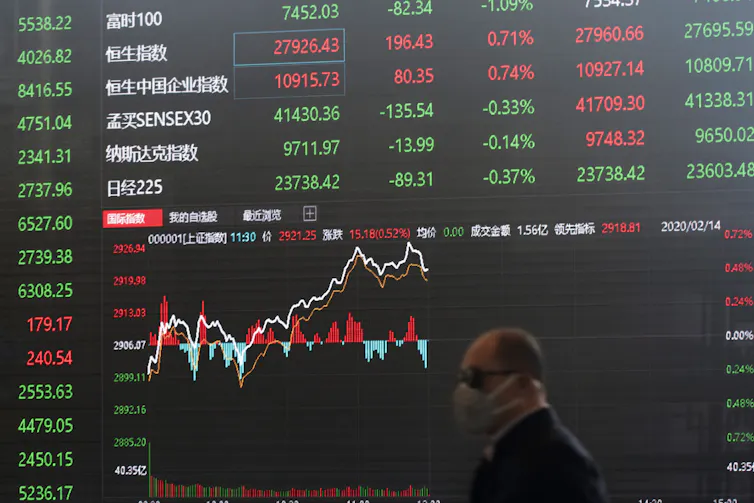 A man in a face mask walks in front of a digital display board showing stock exchange numbers