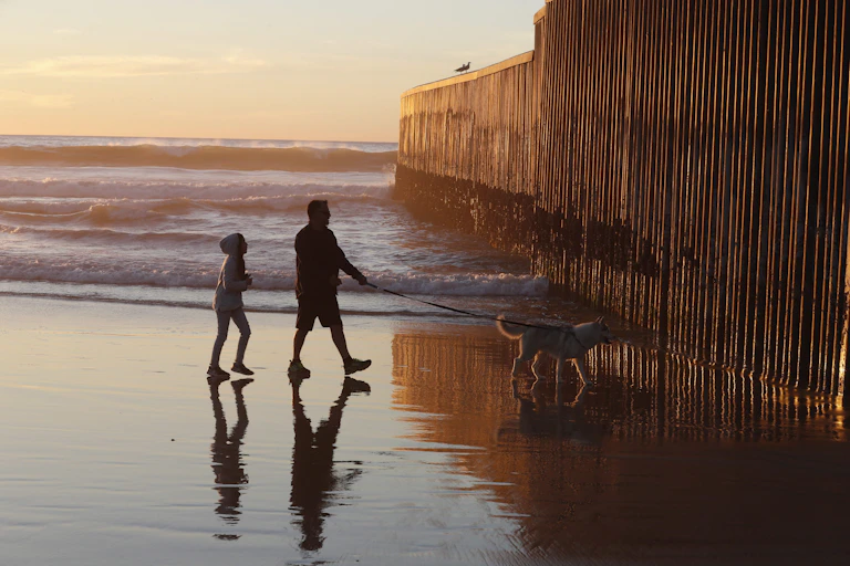 A man and a child and a dog on a leash approach an imposing wall on a beach at sunset.