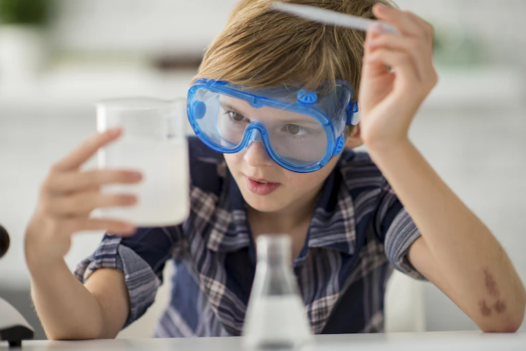 A Caucasian elementary school girl is indoors in a science lab. She is wearing casual clothing and goggles. She is doing a chemistry experiment with a liquid in a container.