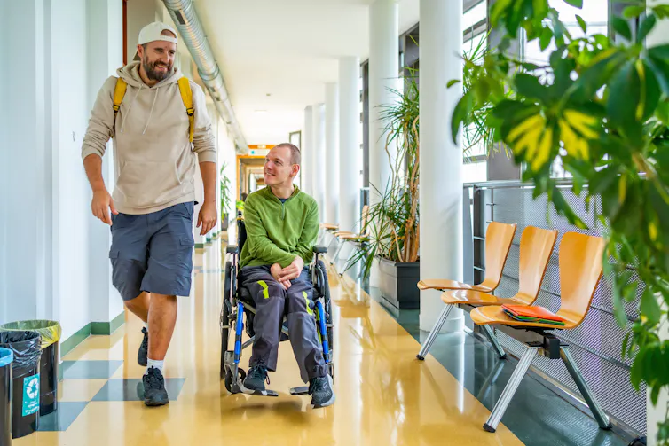 Two smiling men in a corridor. One is using a wheelchair.