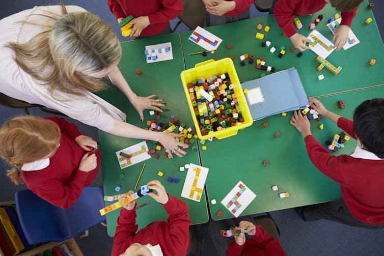 A group of young students and a teacher work with counters at a communal desk.