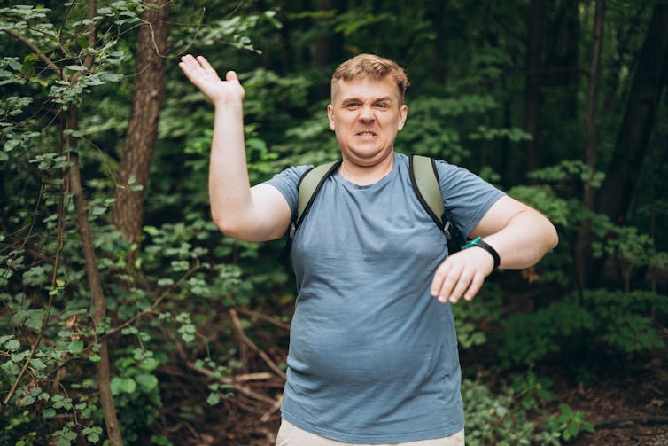 White man walking in a forest waving away mosquitoes.