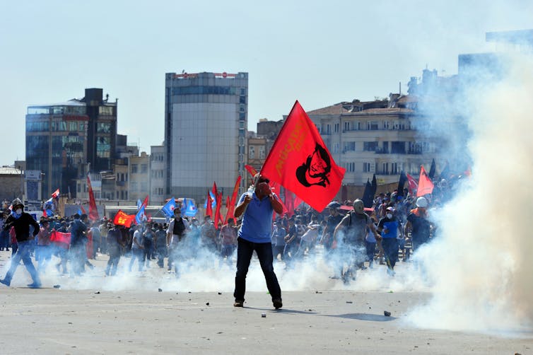 A Turkish protester holding a red flag and covering his face surrounded by smoke.
