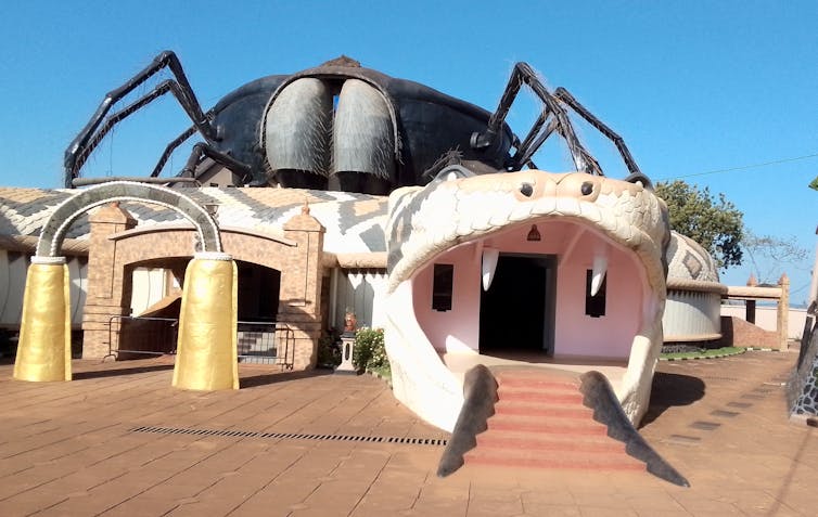 A building with a giant two-headed snake, its mouths serving as entrances and exits with stairs, a giant spider perched on top of it.