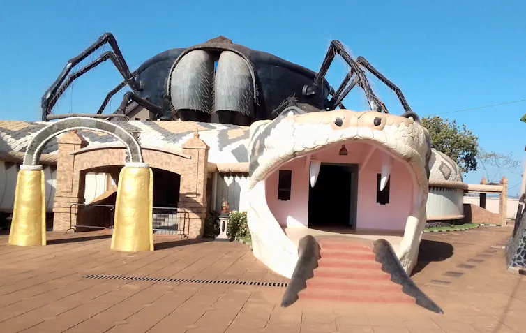 A building with a giant two-headed snake, its mouths serving as entrances and exits with stairs, a giant spider perched on top of it.
