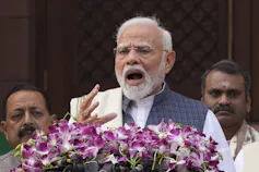 A man with white hair and a white beard speaks at a podium covered with pink flowers.