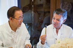 Two men wearing white shirts sit next to each other. One is holding a pen made from a recycled bullet casing.