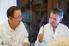 Two men wearing white shirts sit next to each other. One is holding a pen made from a recycled bullet casing.