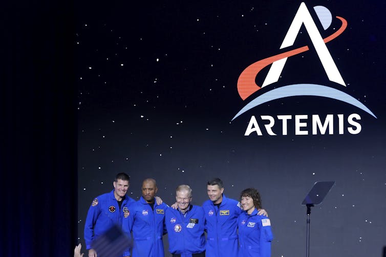 Five people in blue jumpsuits in front of a screen showing the Artemis logo.