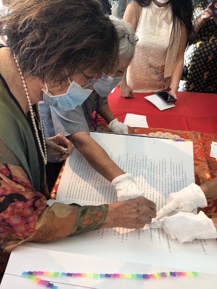 A person's hands hold a small stamp while other, gloved, hands point to a specific area on a page in a large book.