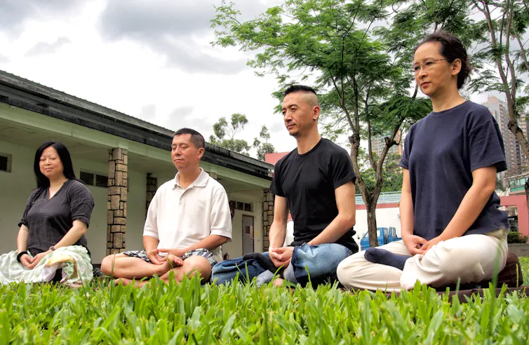 Four individuals seated in a meditation pose on a grassy patch of land.