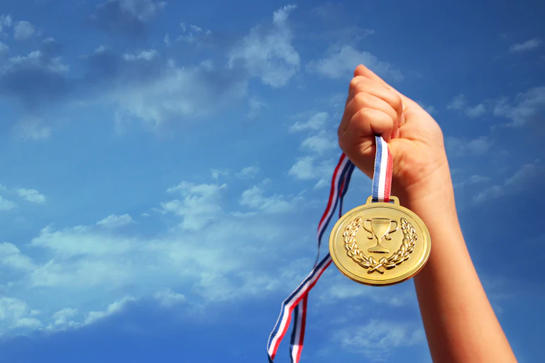 A child's hand holds a gold medal against a blue sky.