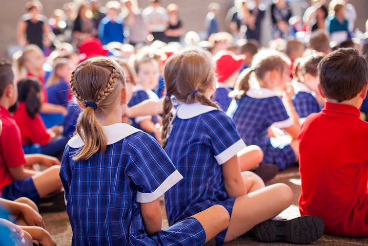 Primary students in uniforms sit on the floor in rows at an assembly.
