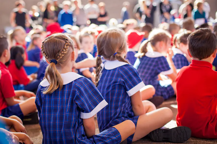 Primary students in uniforms sit on the floor in rows at an assembly.