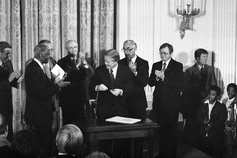 Men in suits surround President Jimmy Carter in a black and white photo.