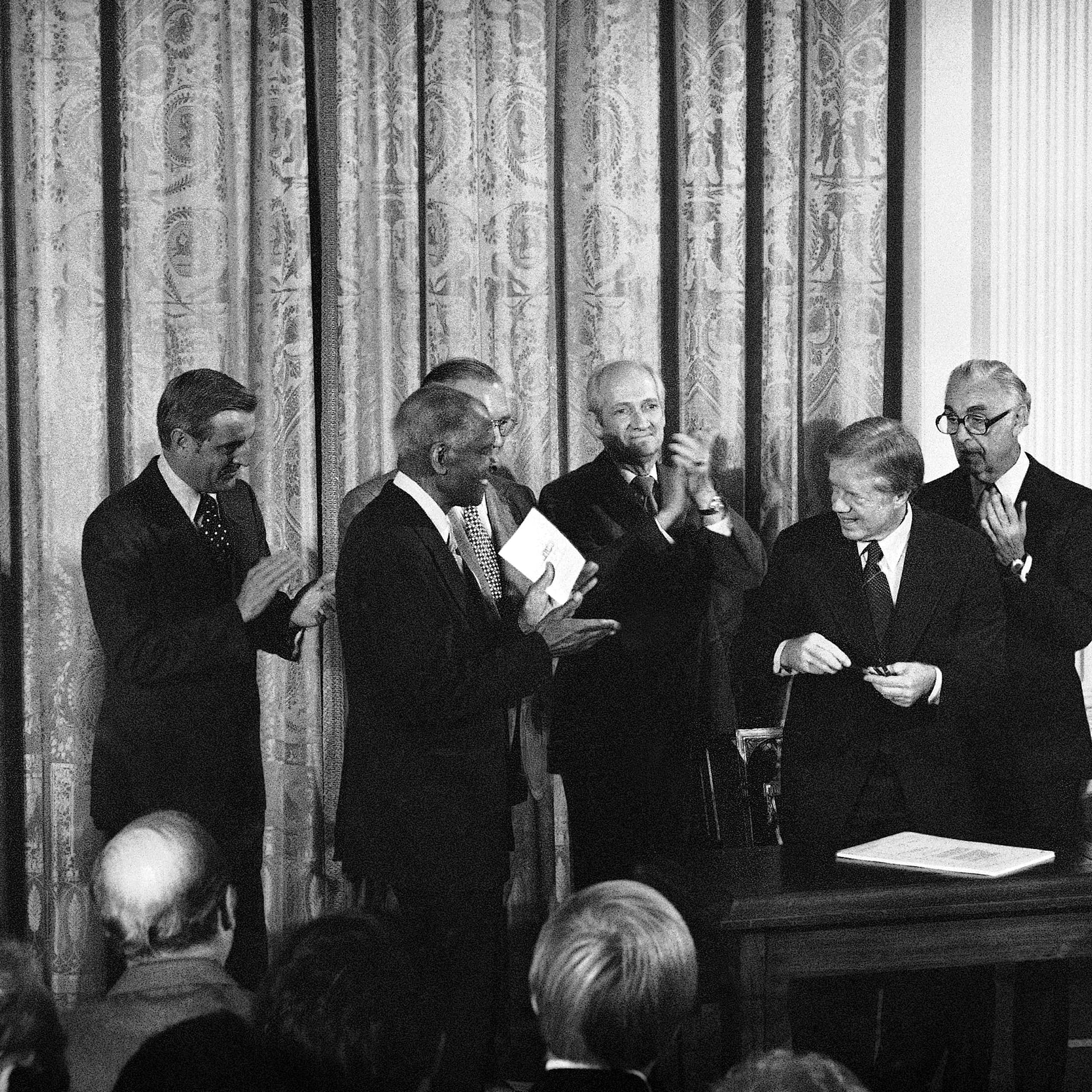 Men in suits surround President Jimmy Carter in a black and white photo.
