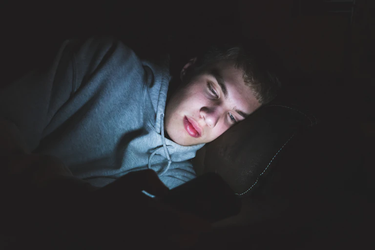 A teenage boy laying in the dark illuminated by his phone